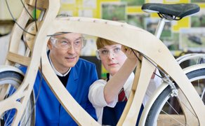 Ein Mann und eine Frau arbeiten in blauer Arbeitskleidung an einem Holzrahmen eines Fahrrads in einer Werkstatt.