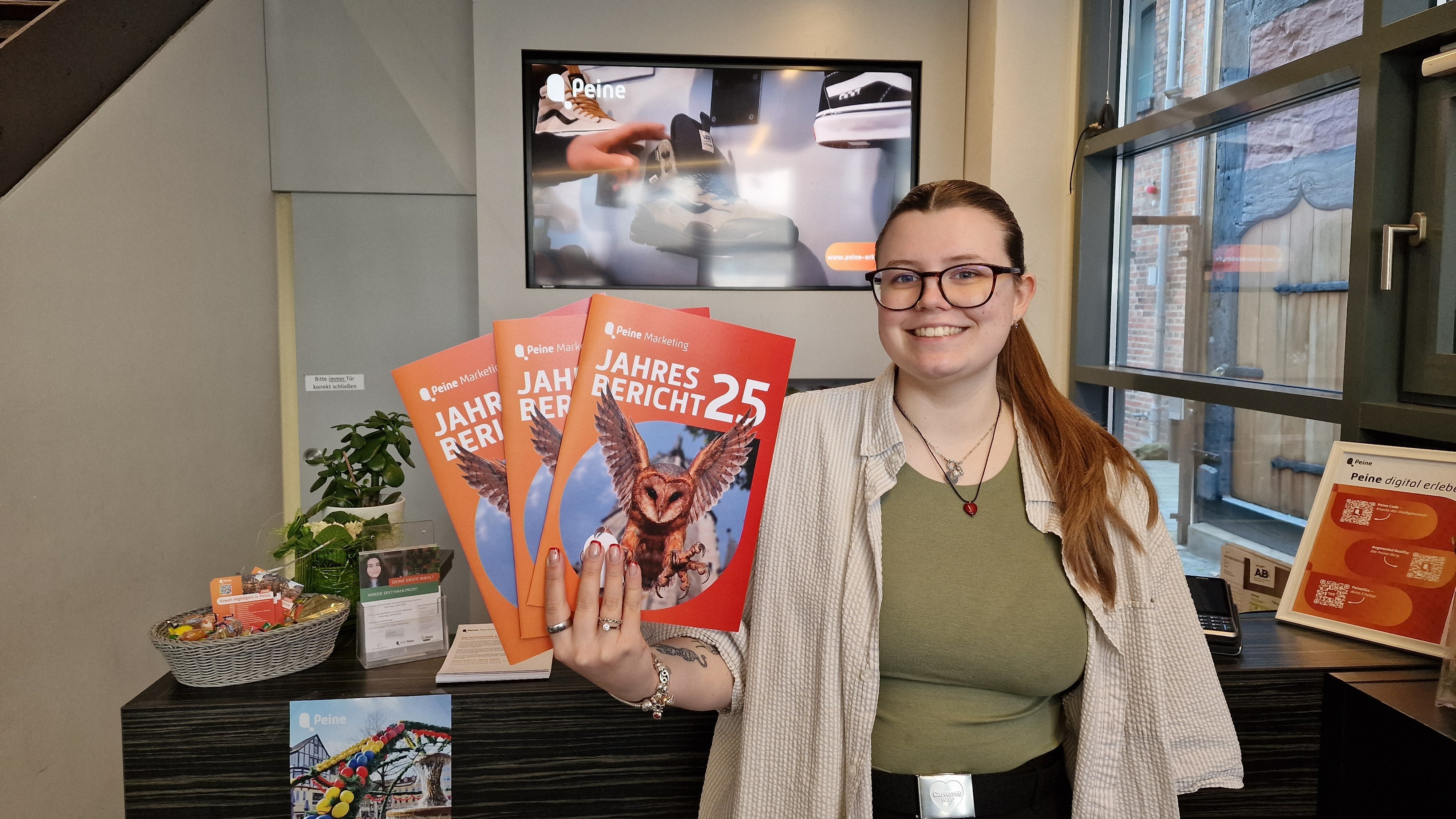 Eine Mitarbeiterin der Peine Marketing GmbH steht in der Tourist-Information und hält mehrere Exemplare des Jahresberichts 2025 in der Hand. Auf den orangefarbenen Titelseiten ist eine fliegende Eule abgebildet.