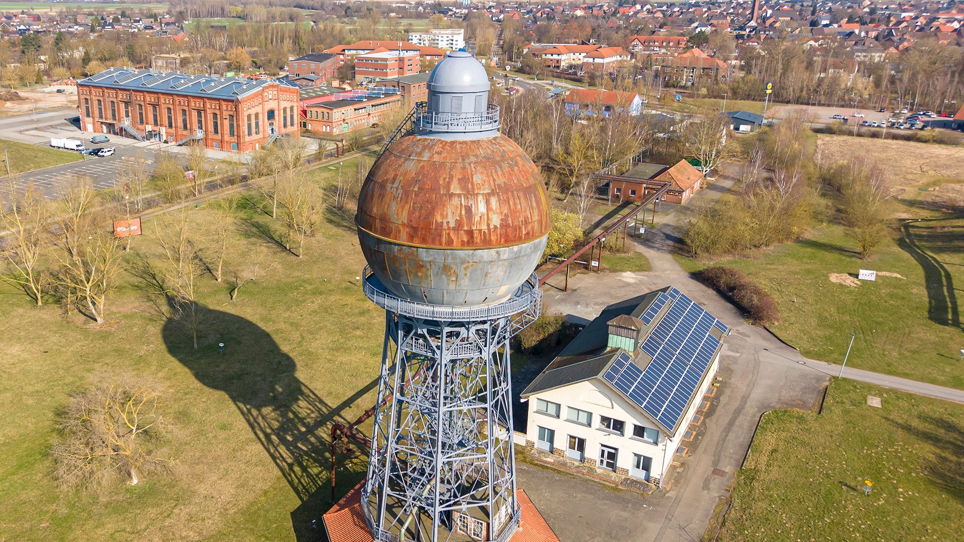 Luftaufnahme eines alten Kugelwasserturms auf einem Industriegelände mit angrenzendem Gebäude und weiteren Industrieanlagen im Hintergrund