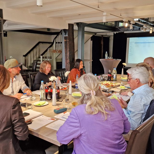 Mehrere Personen sitzen gemeinsam an einem großen Tisch in einem Veranstaltungsraum. Vor ihnen liegen Unterlagen und Notizen, auf dem Tisch stehen Getränke und kleine Snacks. Im Hintergrund ist eine Präsentationsfläche zu sehen.