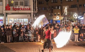 Feuerspucker in rotem Oberteil und schwarzem Rock führt abends vor einer Menschenmenge auf gepflastertem Platz eine Feuershow auf, im Hintergrund ein Gebäude mit Sparkassen-Logo.