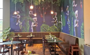 Innenraum eines Cafés mit Sitzbänken und Tischen, dekoriert mit einer großflächigen Wandillustration von Figuren in Kochkleidung zwischen Pflanzen, beleuchtet durch Pendelleuchten.