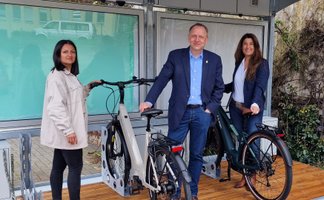 Das Bild zeigt drei Menschen, die mit Fahrrädern an einem BikePort stehen. 