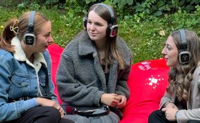 Das Bild zeigt drei Frauen, die sich unterhalten und dabei Kopfhörer auf haben. 