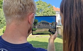Zwei Personen betrachten ein Tablet, auf dem eine digitale Rekonstruktion eines historischen Gebäudes über der heutigen Parklandschaft eingeblendet ist. Im Hintergrund sind Rasenflächen, Bäume und Gebäude zu sehen.