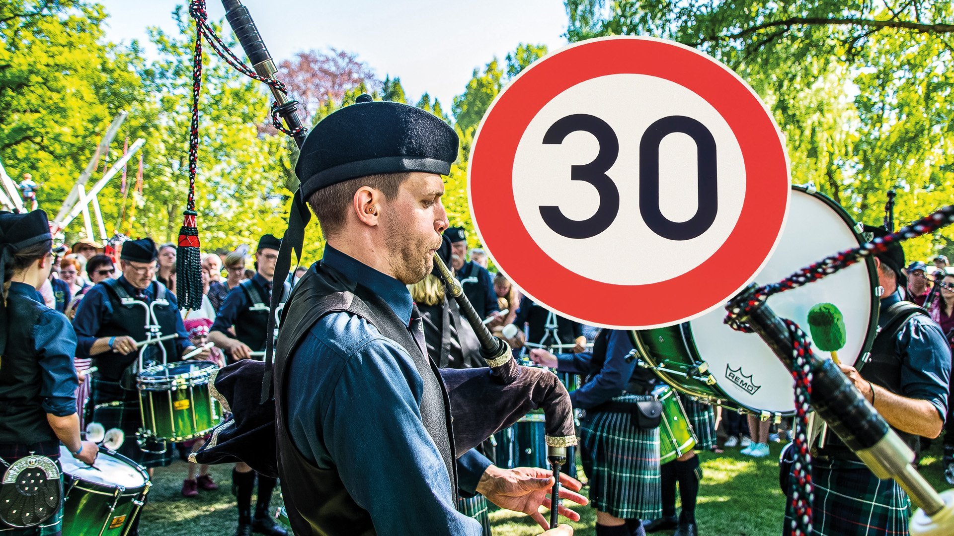 Ein Dudelsackspieler in traditioneller schottischer Kleidung spielt bei einem Highland Gathering, umgeben von einer Trommlergruppe und Zuschauern im Freien; im Vordergrund ist ein rundes Tempo-30-Verkehrsschild zu sehen.
