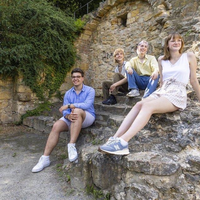 Vier junge Menschen sitzen auf alten Steinmauern und Treppenstufen in einer historischen Ruine mit Bäumen im Hintergrund.