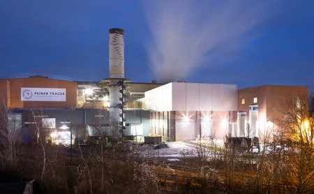 Industrieanlage der Peiner Träger bei Nacht mit beleuchteten Hallen, Schornstein und aufsteigendem Dampf vor blauem Abendhimmel.