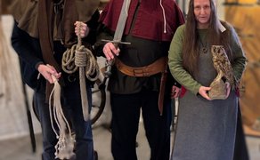 Drei Personen in historischen Kostümen stehen in einem Innenraum und präsentieren Gegenstände wie Seil, Schwert und Eulenfigur; die Szene erinnert an eine Ausstellung zur mittelalterlichen Justiz und Hexenverfolgung.