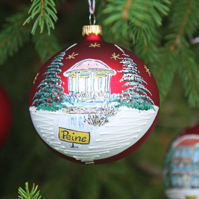 Rote Christbaumkugel mit winterlicher Landschaft und Schriftzug 'Peine' hängt an Tannenzweig neben einer einfarbigen roten Kugel.