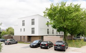 Modernes Wohngebäude mit weißer Fassade und braunem Sockel, davor mehrere geparkte Autos und ein großer Baum.