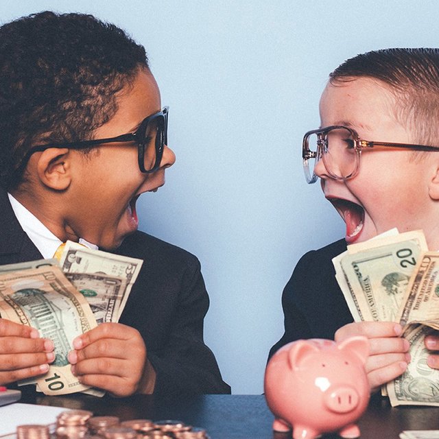 Zwei Jungen in Anzügen sitzen an einem Tisch und halten Geldscheine, auf dem Tisch liegen Münzen, ein Taschenrechner und ein Sparschwein.