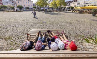 Mehrere Kinder liegen nebeneinander auf einer Holzbank und blicken entspannt in den Himmel. Die Bank steht auf einem großen, gepflasterten Marktplatz, umgeben von historischen Gebäuden, Bäumen und Außengastronomie. Im Hintergrund sind einzelne Passanten zu sehen. Die Szene vermittelt eine ruhige Pause während einer Stadtführung in der Innenstadt.