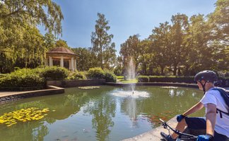 Ein Mann mit Fahrradhelm sitzt am Rand eines Teichs in einem gepflegten Park und blickt auf das Wasser mit einer kleinen Fontäne. Im Teich schwimmen Fische und Seerosenblätter, umgeben von grünen Bäumen und Sträuchern. Im Hintergrund steht ein runder Pavillon. Die Szene zeigt eine ruhige, sonnige Umgebung und vermittelt eine aktive Pause während einer Radtour.