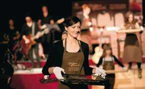 Frau mit Schürze und weißen Handschuhen trägt Tablett mit Schokoladenstücken vor einer Bühne mit Musikern.