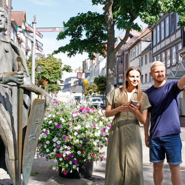 Zwei Teilnehmende stehen neben einer Statue in der Innenstadt von Peine und orientieren sich per Smartphone während einer digitalen Stadtrallye.