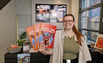 Eine Mitarbeiterin der Peine Marketing GmbH steht in der Tourist-Information und hält mehrere Exemplare des Jahresberichts 2025 in der Hand. Auf den orangefarbenen Titelseiten ist eine fliegende Eule abgebildet.