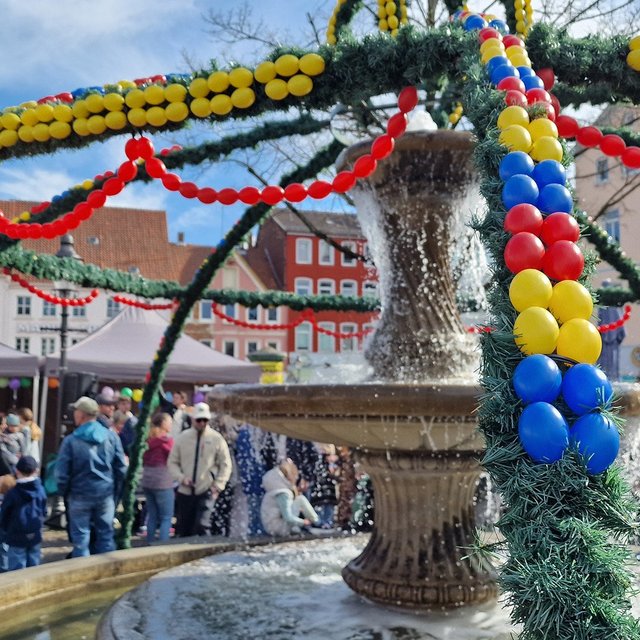 Der Osterbrunnen auf dem Peiner Marktplatz ist mit grünen Girlanden und bunten Ostereiern geschmückt, während sich viele Menschen rund um den Brunnen versammeln.