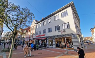 Mehrstöckiges Geschäftshaus an einer belebten Fußgängerzone mit mehreren Passanten und Bäumen.