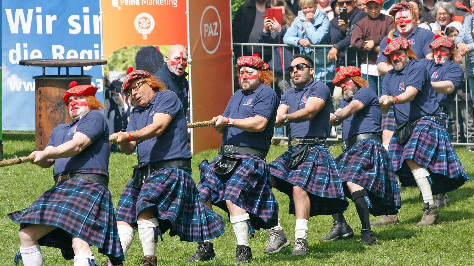 Gruppe von Männern in Kilts und karierten Mützen mit Bommel zieht gemeinsam an einem Tau im Peiner Stadtpark.