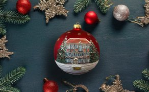 Rote Weihnachtskugel mit bemaltem Haus und Tannenbäumen, umgeben von Tannenzweigen und weiteren roten und silbernen Kugeln.