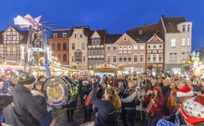 Menschenmenge auf einem Weihnachtsmarkt vor historischen Fachwerkhäusern, mit einer Blaskapelle und einem beleuchteten Weihnachtskarussell im Hintergrund.