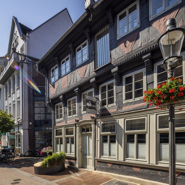 Historisches Fachwerkhaus in der Peiner Innenstadt mit liebevollen Details: helle Fensterrahmen, Blumen am Laternenmast und eine ruhige Fußgängerzone mit Cafés und Fahrrädern – ein stimmungsvolles Beispiel für den kulturellen Treffpunkt rund um den Kulturschwan in Peine.