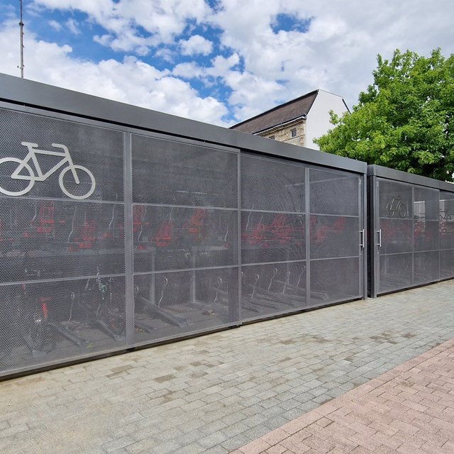 Überdachte Bike-and-Ride-Fahrradboxen in der Innenstadt von Peine mit verschließbaren Abteilen und Fahrrad-Symbol, auf einem gepflasterten Gehweg neben Bäumen und Gebäuden.
