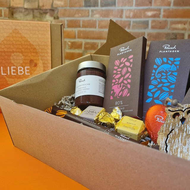 Offene Genussbox mit drei Rausch Plantagen Schokoladentafeln, einem Glas Marmelade, mehreren Pralinen und einer Holz-Eulenfigur vor Ziegelwand.