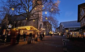 Weihnachtlich beleuchteter Kirchplatz mit Fachwerkhäusern, Baum mit Lichterkugeln und Menschen bei Dämmerung.