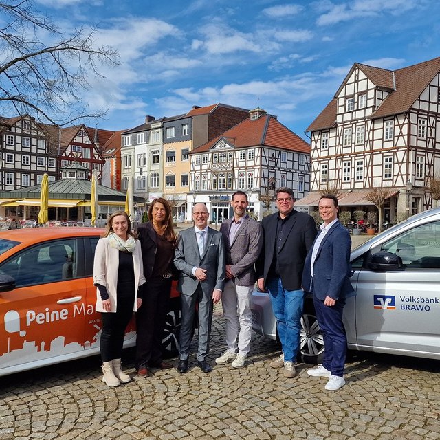Sechs Personen stehen zwischen einem orangefarbenen Auto mit der Aufschrift „Peine“ und einem silbernen Fahrzeug mit dem Logo „Volksbank BRAWO“ vor Fachwerkhäusern auf einem gepflasterten Platz.