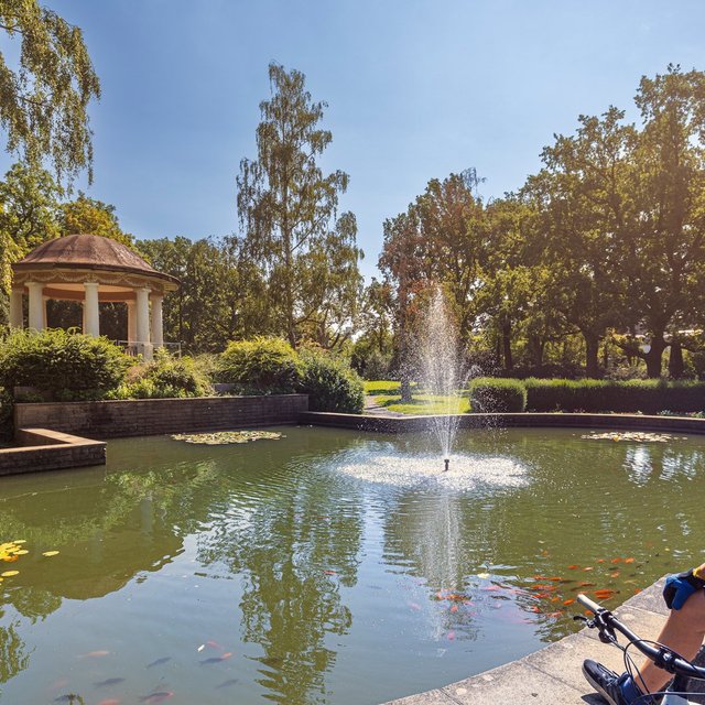 Ein Mann mit Fahrradhelm sitzt am Rand eines Teichs in einem gepflegten Park und blickt auf das Wasser mit einer kleinen Fontäne. Im Teich schwimmen Fische und Seerosenblätter, umgeben von grünen Bäumen und Sträuchern. Im Hintergrund steht ein runder Pavillon. Die Szene zeigt eine ruhige, sonnige Umgebung und vermittelt eine aktive Pause während einer Radtour.