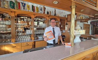 Ein Mann steht hinter einer Bar und hält ein orangefarbenes Schild mit der Aufschrift „HIER kostenlos“.