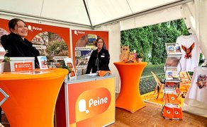 Messestand von Peine Marketing beim Entdeckertag mit orangefarbenen Infoständen, Informationsmaterialien und zwei Mitarbeiterinnen.