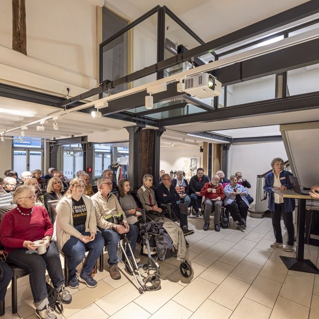 Eine Gruppe von rund 30 überwiegend älteren Menschen sitzt in einem modernisierten Fachwerkraum und hört aufmerksam einer Referentin zu, die rechts neben einer Präsentationsfläche steht und einen Vortrag hält.