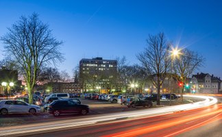 Abendliche Ansicht eines Parkplatzes in der Peiner Innenstadt mit beleuchteten Straßen, parkenden Autos und Lichtspuren des vorbeifahrenden Verkehrs.