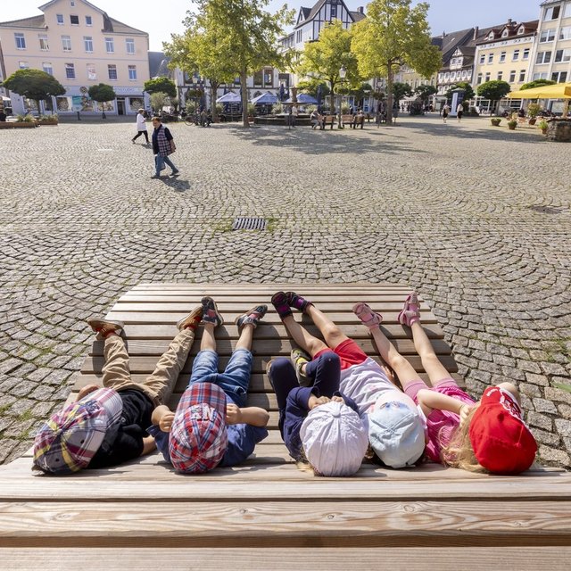 Mehrere Kinder liegen nebeneinander auf einer Holzbank und blicken entspannt in den Himmel. Die Bank steht auf einem großen, gepflasterten Marktplatz, umgeben von historischen Gebäuden, Bäumen und Außengastronomie. Im Hintergrund sind einzelne Passanten zu sehen. Die Szene vermittelt eine ruhige Pause während einer Stadtführung in der Innenstadt.
