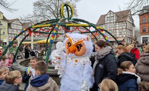 Eine als Eule verkleidete Person steht vor einem geschmückten Brunnen auf einem Marktplatz und winkt. Rundherum befinden sich viele Kinder und Erwachsene, einige schauen zur Figur, andere unterhalten sich. Der Brunnen ist mit grünen Girlanden und bunten Ostereiern dekoriert.