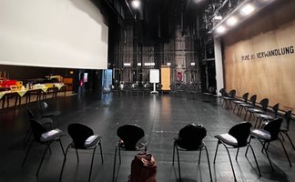 Das Bild zeigt eine Theaterbühne, auf der sich zahlreiche leere Stühle befinden, die im Kreis aufgestellt sind. 