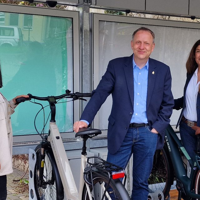 Das Bild zeigt drei Menschen, die mit Fahrrädern an einem BikePort stehen. 