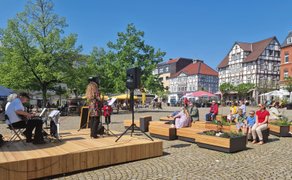 Open-Stage-Veranstaltung auf dem Marktplatz in Peine: Musikerinnen und Musiker treten auf einer kleinen Holz-Bühne auf, während Besucherinnen und Besucher bei sonnigem Wetter auf Sitzbänken die Live-Musik und die historische Kulisse der Innenstadt genießen.