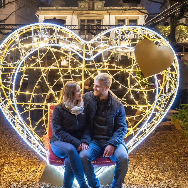 Das Bild zeigt ein Pärchen, das sich anschaut und dabei im beleuchteten Herz sitzt. Einer XXL-Lichtfigur in der Weihnachtsstadt Peine.