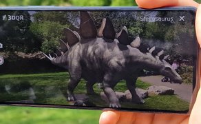 Augmented-Reality-Darstellung eines Stegosaurus auf dem Display eines Smartphones in einer Parklandschaft.