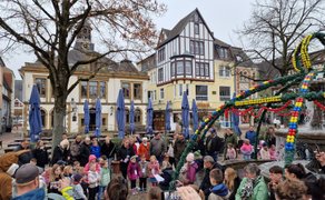 Viele Menschen, darunter zahlreiche Kinder und Erwachsene, stehen auf einem Marktplatz um einen geschmückten Brunnen herum. Der Brunnen ist mit grünen Girlanden und bunten Ostereiern dekoriert. Einige Kinder stehen im Mittelpunkt und hören aufmerksam zu, während Erwachsene zuschauen und teilweise Fotos machen. Im Hintergrund sind historische Gebäude, Bäume und geschlossene Sonnenschirme eines Cafés zu sehen.