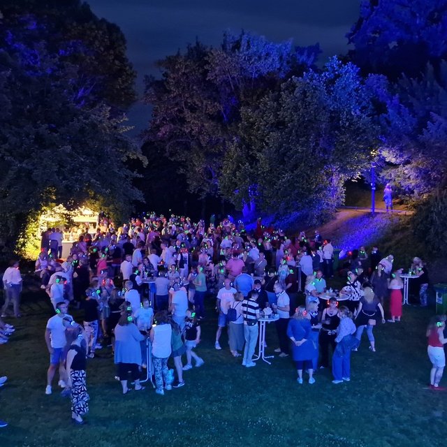 Große Menschenmenge feiert eine Silent Disco im Park bei Nacht, mit leuchtenden Funkkopfhörern, Stehtischen und farbig beleuchteten Bäumen rund um das Veranstaltungsgelände in Peine.