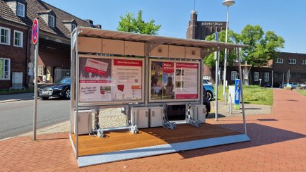 Überdachte Bikeport-Anlage in der Peiner Innenstadt mit Infotafeln, Fahrradboxen und Lademöglichkeiten, daneben E-Ladesäulen und historische Backsteingebäude im Hintergrund.