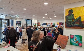 Menschenmenge in einer Galerie mit zahlreichen farbigen Gemälden an den Wänden, darunter ein großes Bild einer sitzenden Figur in Grün auf gelbem Hintergrund.