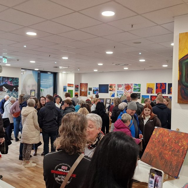 Menschenmenge in einer Galerie mit zahlreichen farbigen Gemälden an den Wänden, darunter ein großes Bild einer sitzenden Figur in Grün auf gelbem Hintergrund.