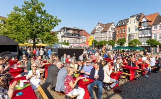 Viele Menschen sitzen an langen, rot gedeckten Tischen auf einem historischen Marktplatz mit Fachwerkhäusern und verfolgen gemeinsam eine Veranstaltung im Freien.