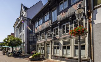 Historisches Fachwerkhaus in der Peiner Innenstadt mit liebevollen Details: helle Fensterrahmen, Blumen am Laternenmast und eine ruhige Fußgängerzone mit Cafés und Fahrrädern – ein stimmungsvolles Beispiel für den kulturellen Treffpunkt rund um den Kulturschwan in Peine.